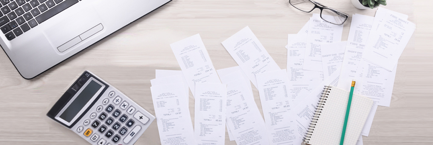 An image of expense bills scattered on the work desk with a calculator, laptop, pencil, and spectacles